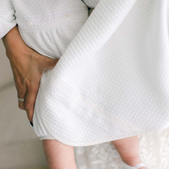A mother covers her baby with the Elijah Newborn Personalized Blanket. The blanket is made from soft plush 100% cotton in white with quilted texture. On the corner is the name "Elijah" embroidered