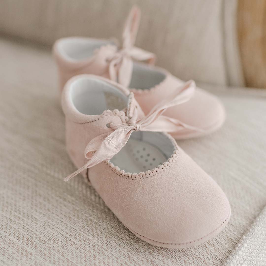 A pair of Joli Suede Tie Mary Janes in soft pink with delicate ribbon ties on a textured beige fabric background, conveying a gentle and cozy upscale aesthetic.