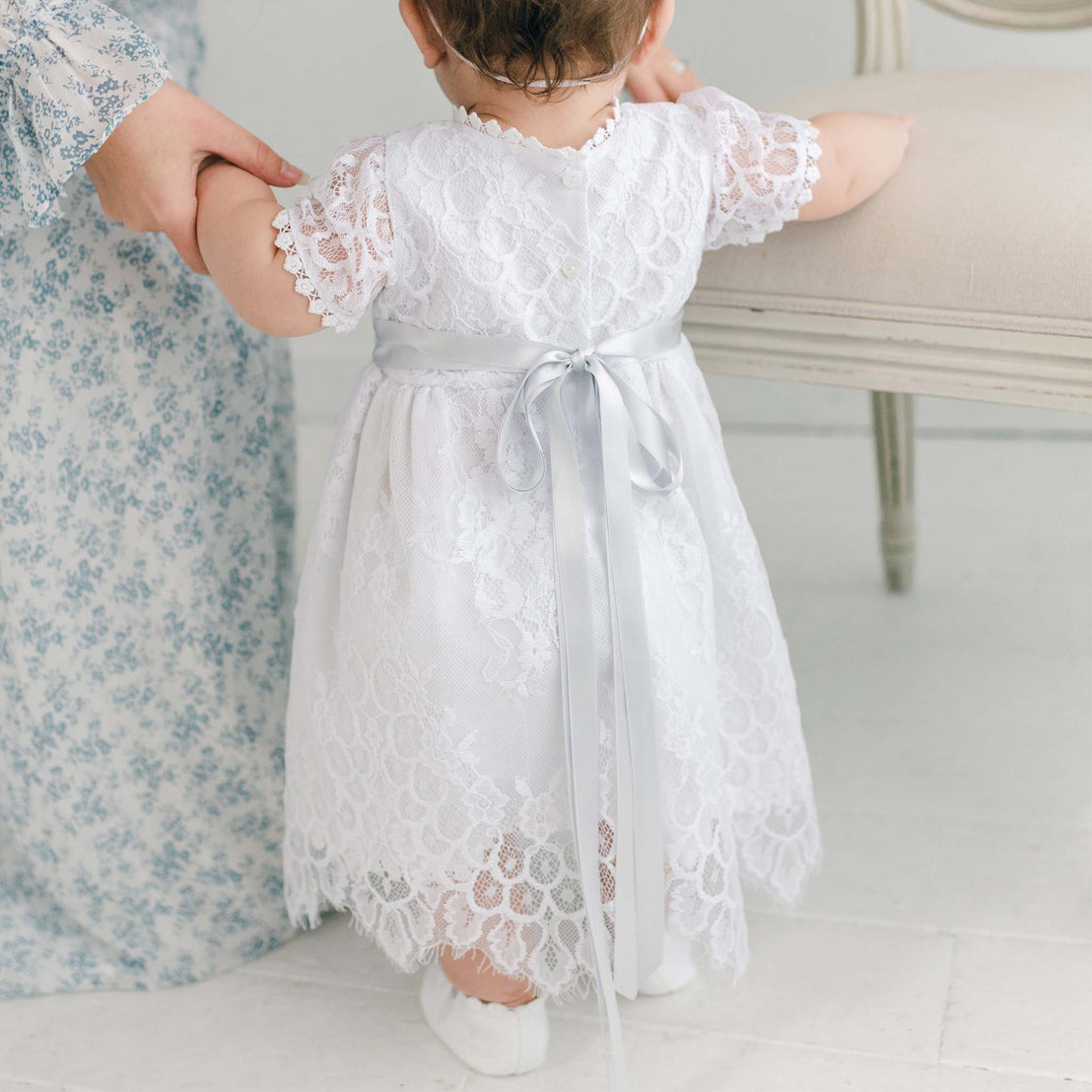 A toddler in an Olivia Dress, learning to stand up with the support of an adult's hands, perfect as a boutique baby gift or for a christening, in a light, airy room