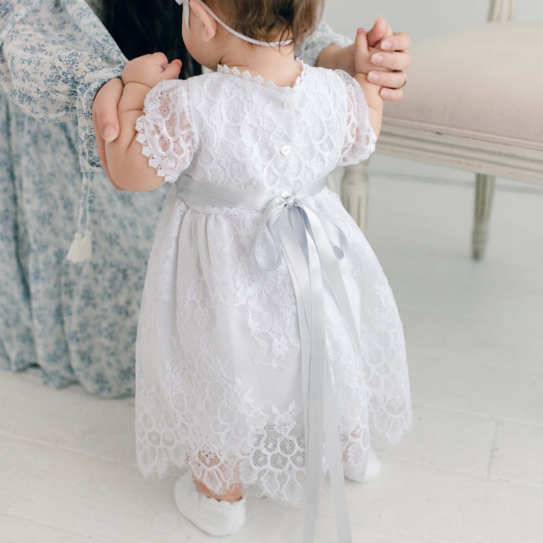 A toddler in an Olivia Dress is held by her mother, both facing away from the camera in a softly lit boutique room.