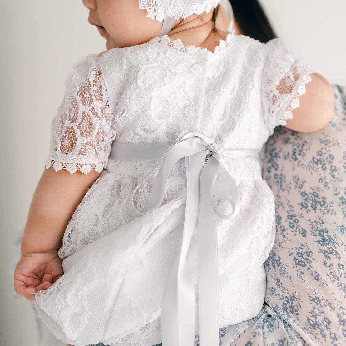 A close-up of a baby dressed in the Olivia Christening Gown & Bonnet, being held securely in a parent's arms, perfect for baptism or as a boutique baby gift.
