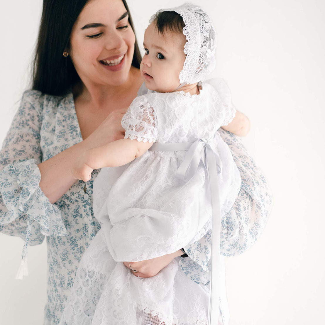 A joyful mother holding her baby girl dressed in the Olivia Christening Gown & Bonnet with lace details, both smiling against a clean, white background.