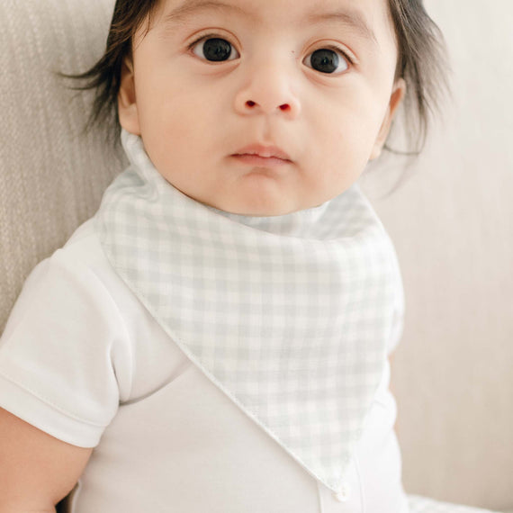 Close-up of a baby with wide eyes, wearing a designer white onesie and an Ian Bandana Bib, lying down on a soft, light-colored surface.