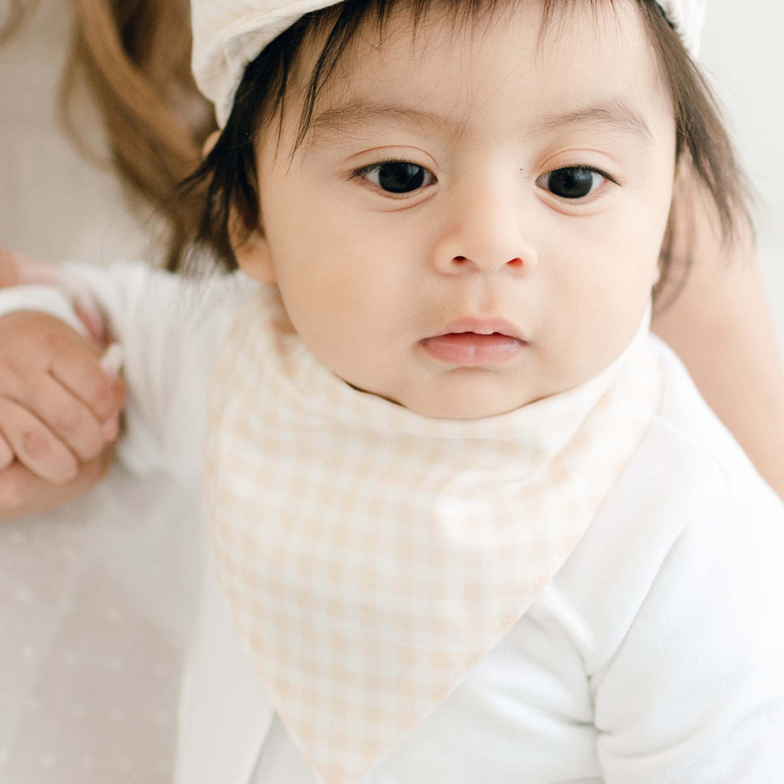 Close-up view of a baby dressed in a designer white outfit with an Ian Bandana Bib and white bonnet, looking directly at the camera with a soft focus background.