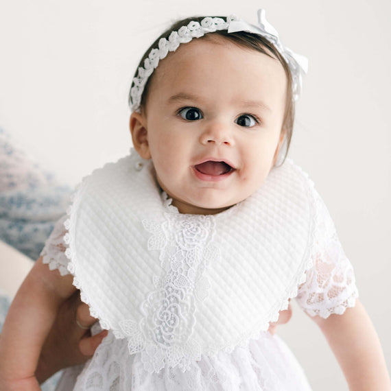 A cheerful baby girl smiles brightly against a soft, neutral background. Her eyes sparkle with joy and curiosity, wearing an Olivia Bib and headband, perfect for a baptism or as a baby.