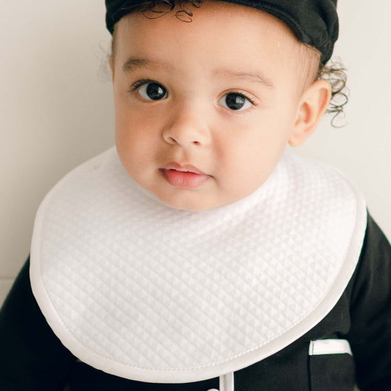Baby boy wearing the James Bib made from 100% white quilted cotton with button closure and silk piping on the edge.