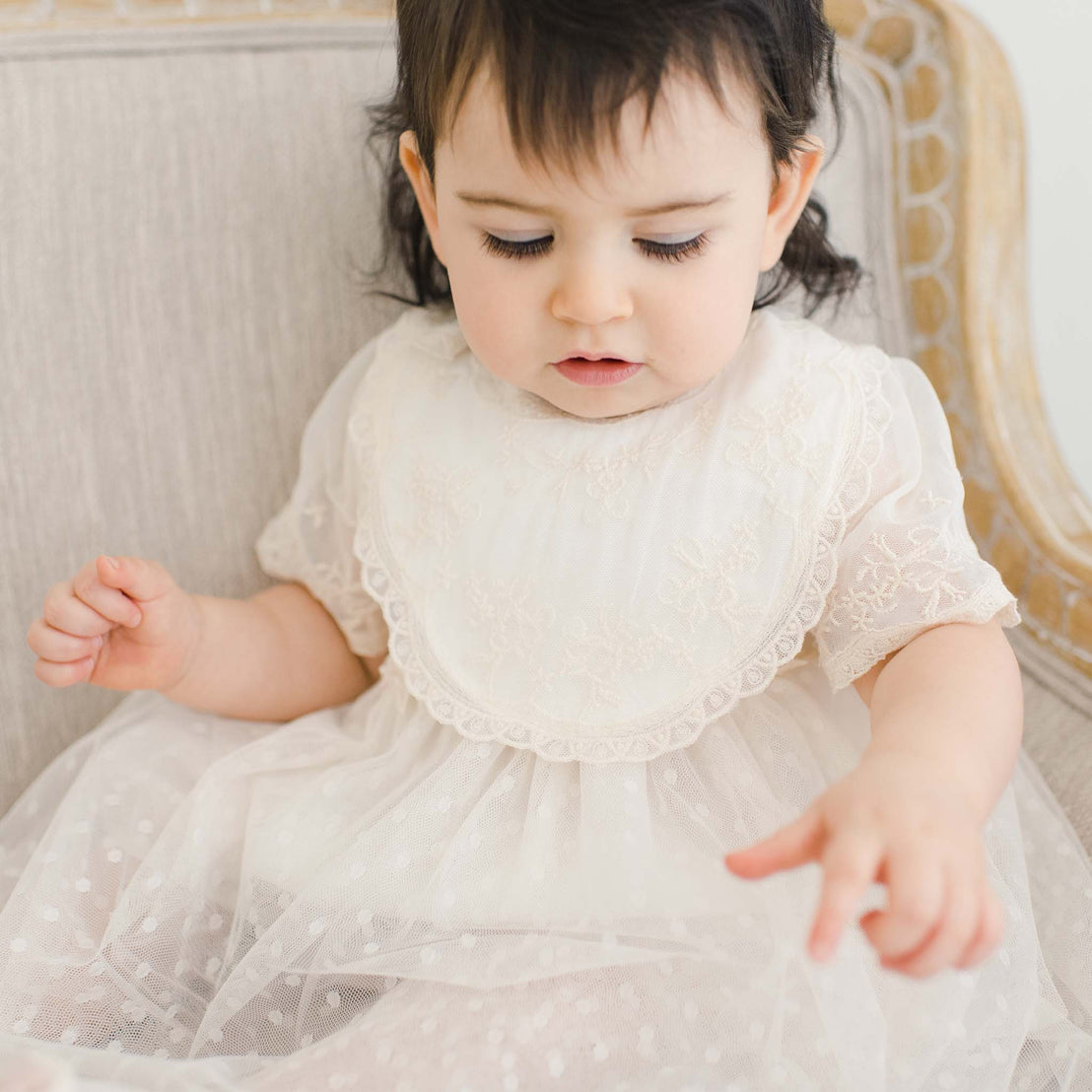 A young toddler with dark hair sits on an elegant cream-colored chair and is wearing the Jessica Cotton Bib, an ivory cotton bib overlayed with an intricate floral embroidery champagne lace, over a lace dress. She looks down curiously.