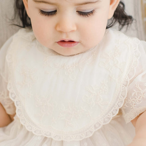 Close-up of a toddler wearing the Jessica Cotton Bib, featuring ivory cotton and a lace overlay in champagne lace, over the Jessica Linen Gown. She is focused intently on something out of view.