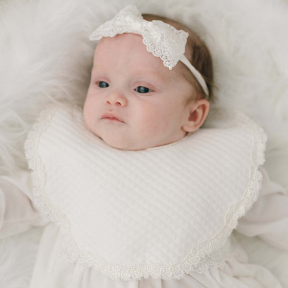 A baby wearing a Madeline Bib and a lace headband, lying on a soft white surface, looking upward with a thoughtful expression.