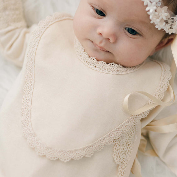 A close-up photo of a baby with blue eyes wearing the Mia Newborn Bib, a soft pima cotton bib in a shade of champagne with cotton lace detailing along the edges, over the Mia Knot Gown.