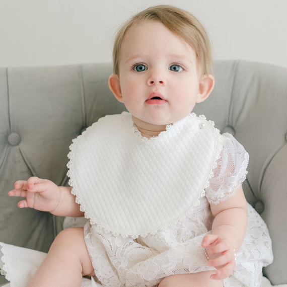 A baby with blue eyes and light brown hair wearing the Aria Bubble Romper and an Aria Bib. She sits on a gray sofa, looking surprised and adorable.