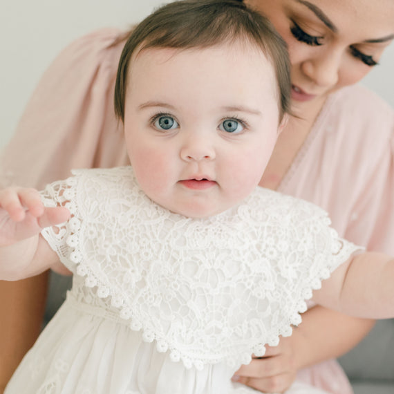 A woman in a soft pink blouse gently holds a baby dressed in a white Christening gown with a large collar. The baby, with striking blue eyes, looks directly at the camera while wearing the Grace Lace Bib.