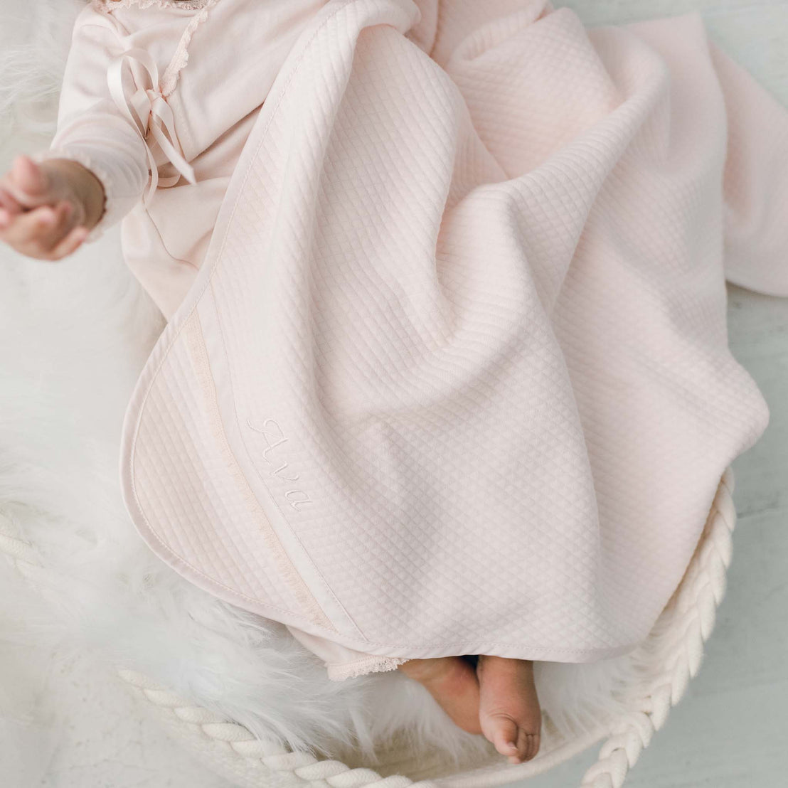 Closeup of a newborn baby, partially covered with the blush pink quilted Ava Personalized Blanket, sits in a woven basket lines with fluffy white fur, and reaches out with one hand.