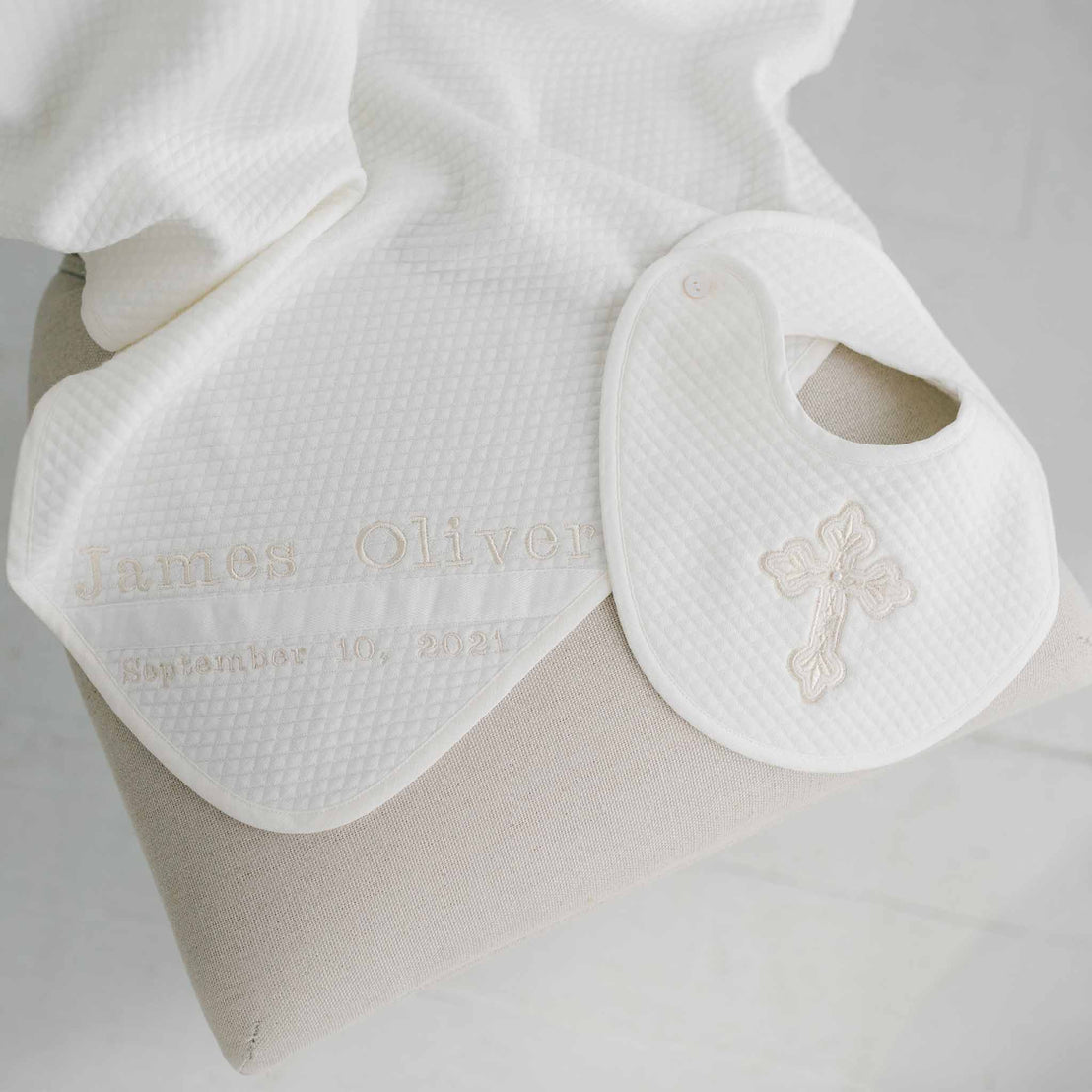 A personalized white Baby Boy Blessing Gift Set bib from Baby Beau & Belle with the name "james oliver" embroidered in light brown thread, alongside a date "10/2021" and a small floral design, resting on a quilted.