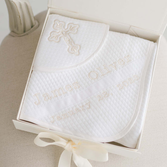 A Baby Beau & Belle Baby Boy Blessing Gift Set, including a personalized white quilted cotton blanket embroidered with the name "James Oliver" and the date "January 23, 2023", displayed in an open gift box with a ribbon.