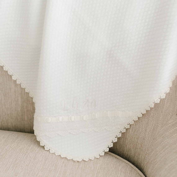 Close-up of a vintage white embroidered Eliza Personalized Blanket with the name "Eliza" stitched in an elegant font, featuring delicate lace trim against a soft fabric background.