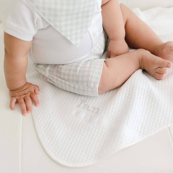 A baby sitting on a soft, white Ian Personalized Blanket with the name "Ian" embroidered on it, showing just the baby's plump legs, hands, and torso dressed in a traditional light blue outfit.