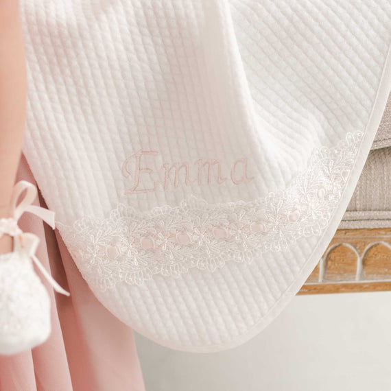 A close-up of a white Emma Personalized Blanket embroidered with the name "Emma" in pink thread, adorned with a delicate lace trim, held by someone wearing a pink dress.