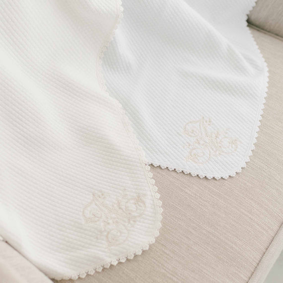 Two white, embroidered  baby blankets with decorative lace edges and textured details, elegantly displayed on a neutral beige surface, featuring letter embroidery personalization.