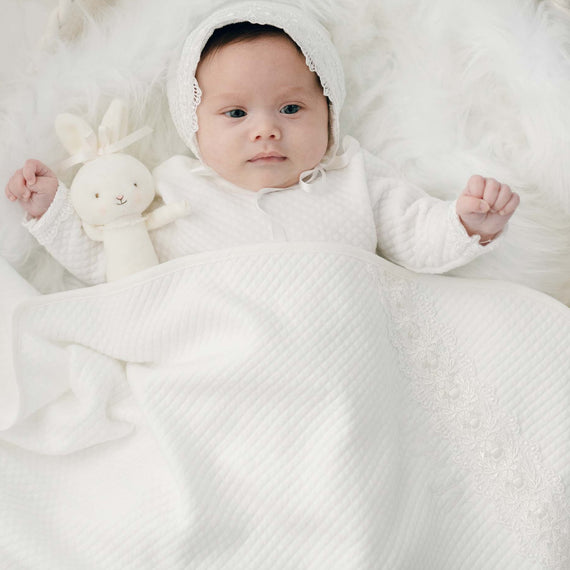 A newborn baby in a white bonnet lies on a fluffy Madeline Personalized Blanket, holding a white bunny toy, covered partially by an heirloom white blanket with lace details.
