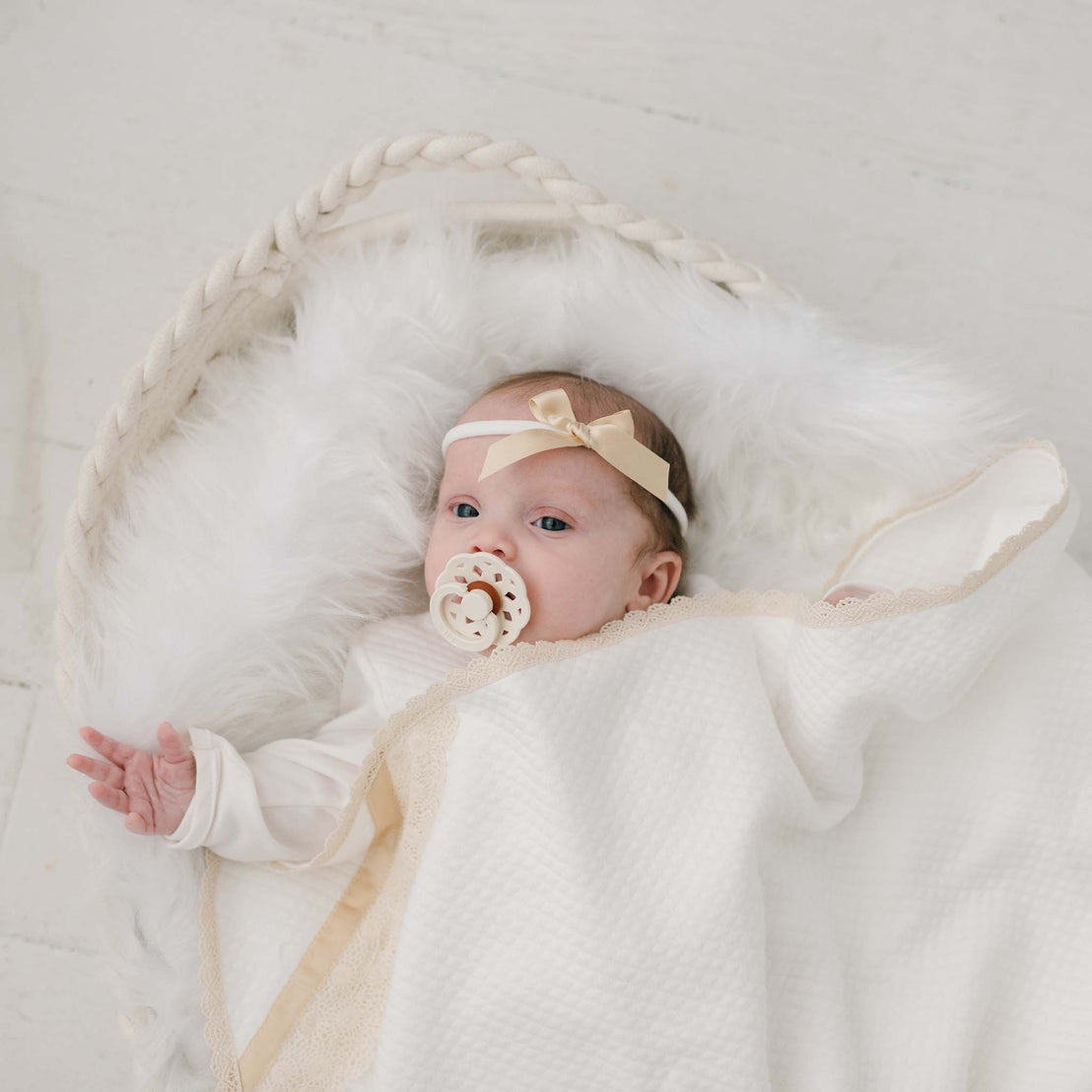 A baby wearing the Mia Bow Headband and using a pacifier lies cozily in a white basket wrapped in the Mia Personalized Blanket.