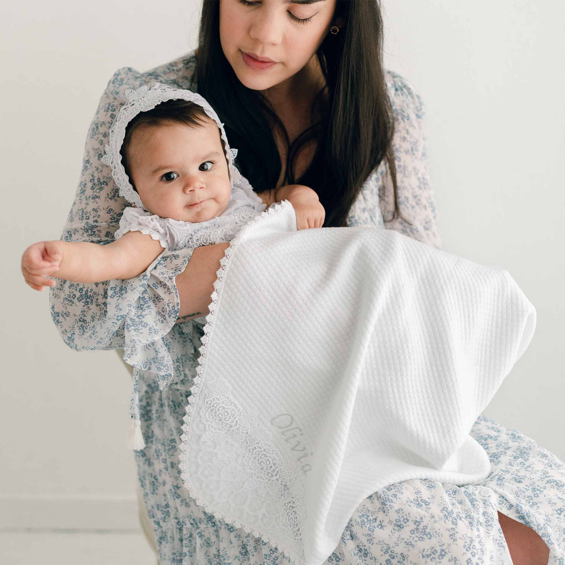 A woman cradles a baby wearing a bonnet, as they both pose for a christening portrait. The baby, dressed in white, looks directly at the camera, while an Olivia Personalized Blanket reading "Olivia" is draped over their laps.