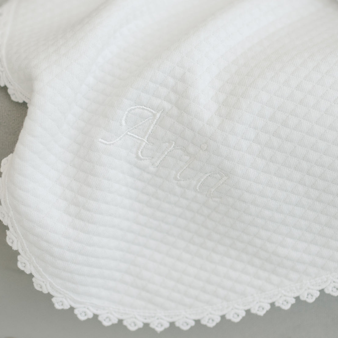 Close-up of a white textured cotton baby Aria Personalized Blanket with the name "Aria" embroidered in white thread, featuring a delicate lace trim.