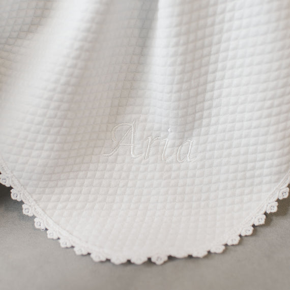 Close-up view of a white textured Aria Personalized Blanket with the name "Aria" embroidered in white thread, detailed with a delicate lace trim along the edge.