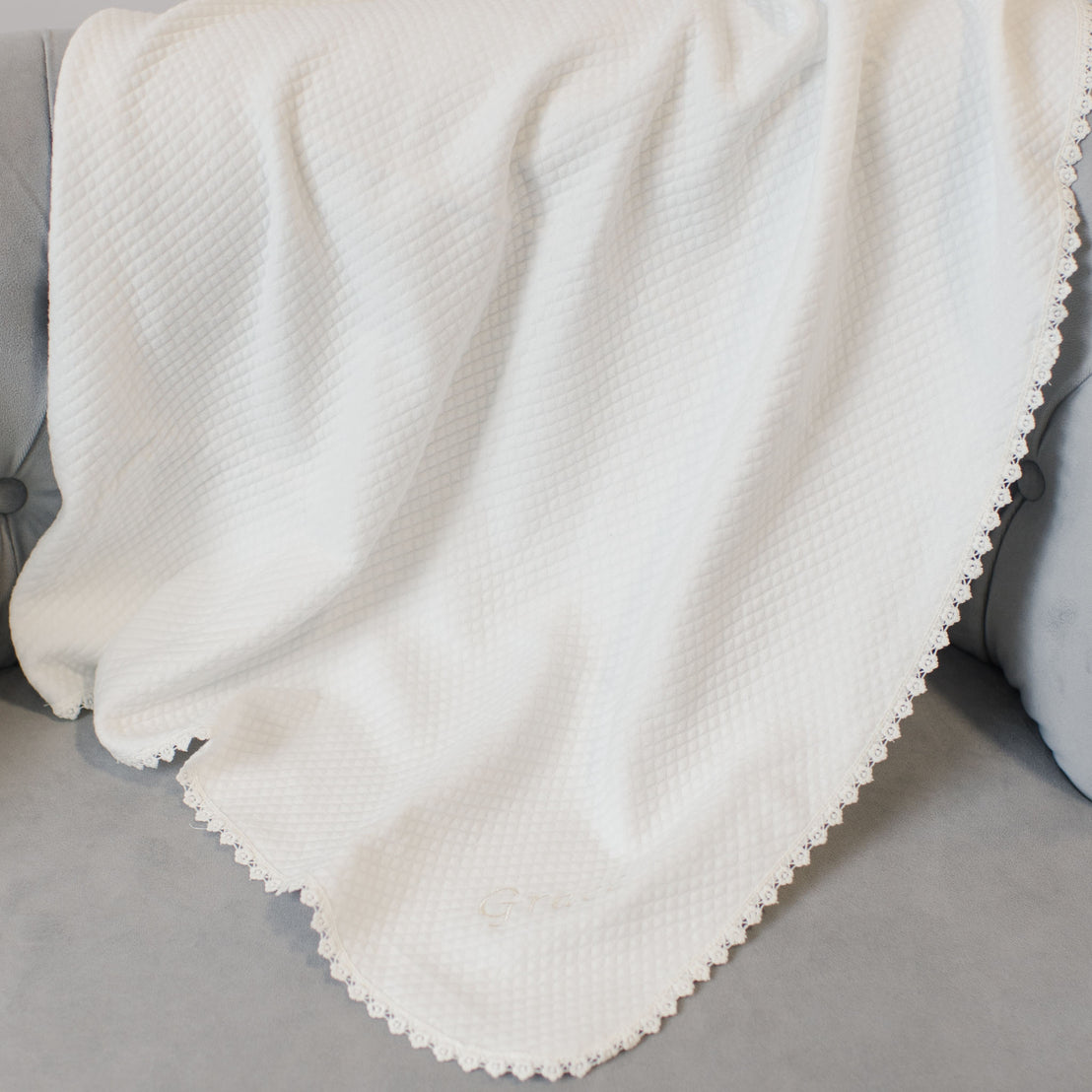 A soft white waffle weave Grace Personalized Blanket with a scalloped edge draped over a gray couch.