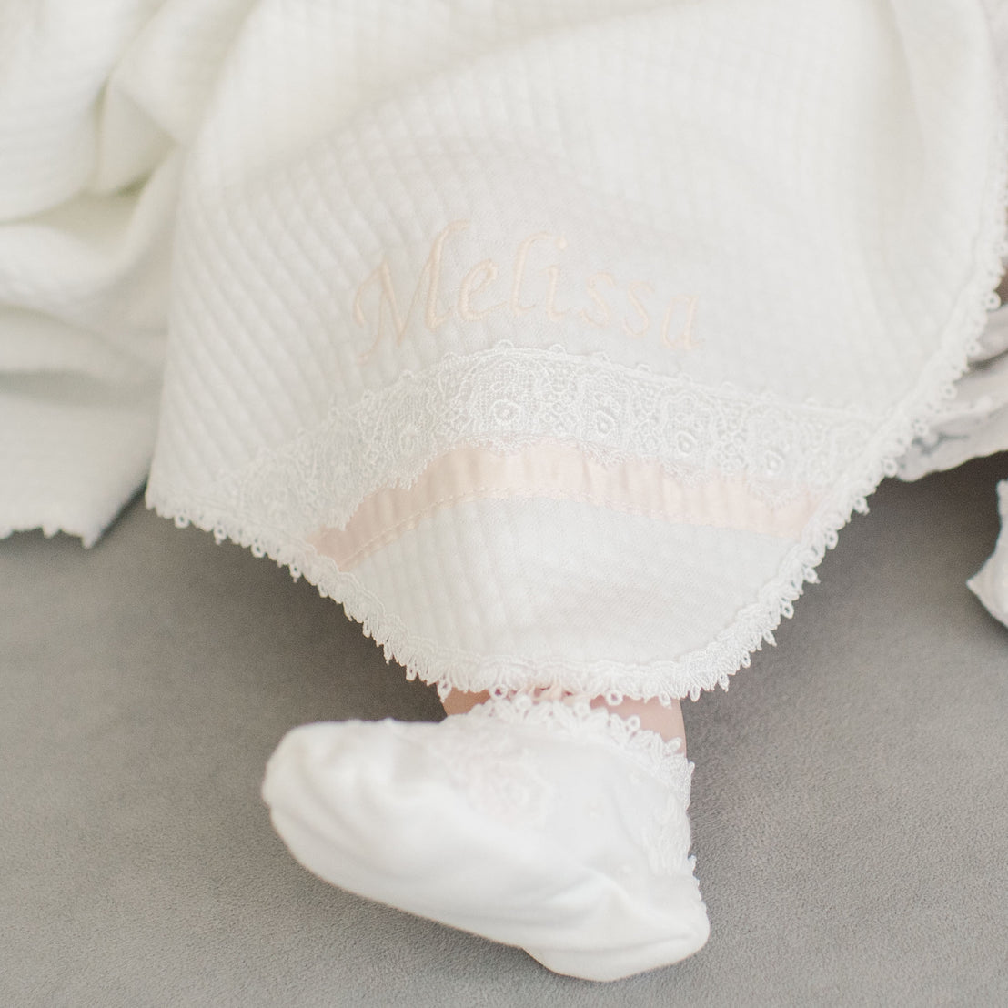 A baby's foot peeks out from under a soft white blanket embroidered with the name "Melissa," surrounded by lacy detailing and is part of the Melissa Accessory Bundle.