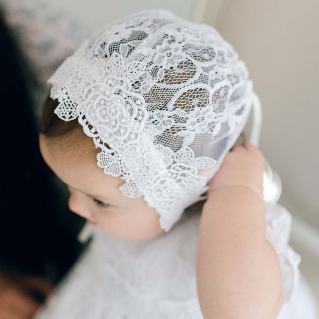 A baby wearing the Olivia Christening Gown & Bonnet looks downward, with a soft focus background enhancing the intricate texture of the lace, perfect for a baptism or as a boutique baby gift.