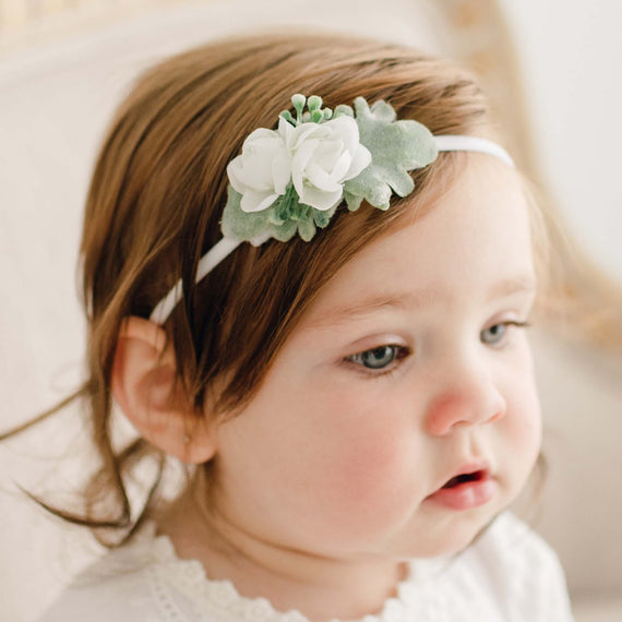 Portrait of a young toddler girl wearing the Eliza Flower Headband, dressed in a white baptism outfit, looking to the side. 