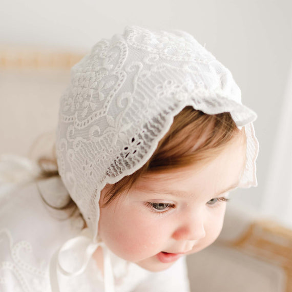 A toddler with light brown hair wears an Eliza Bonnet suitable for a christening and gazes downward with a gentle expression, set against a soft, neutral vintage background.