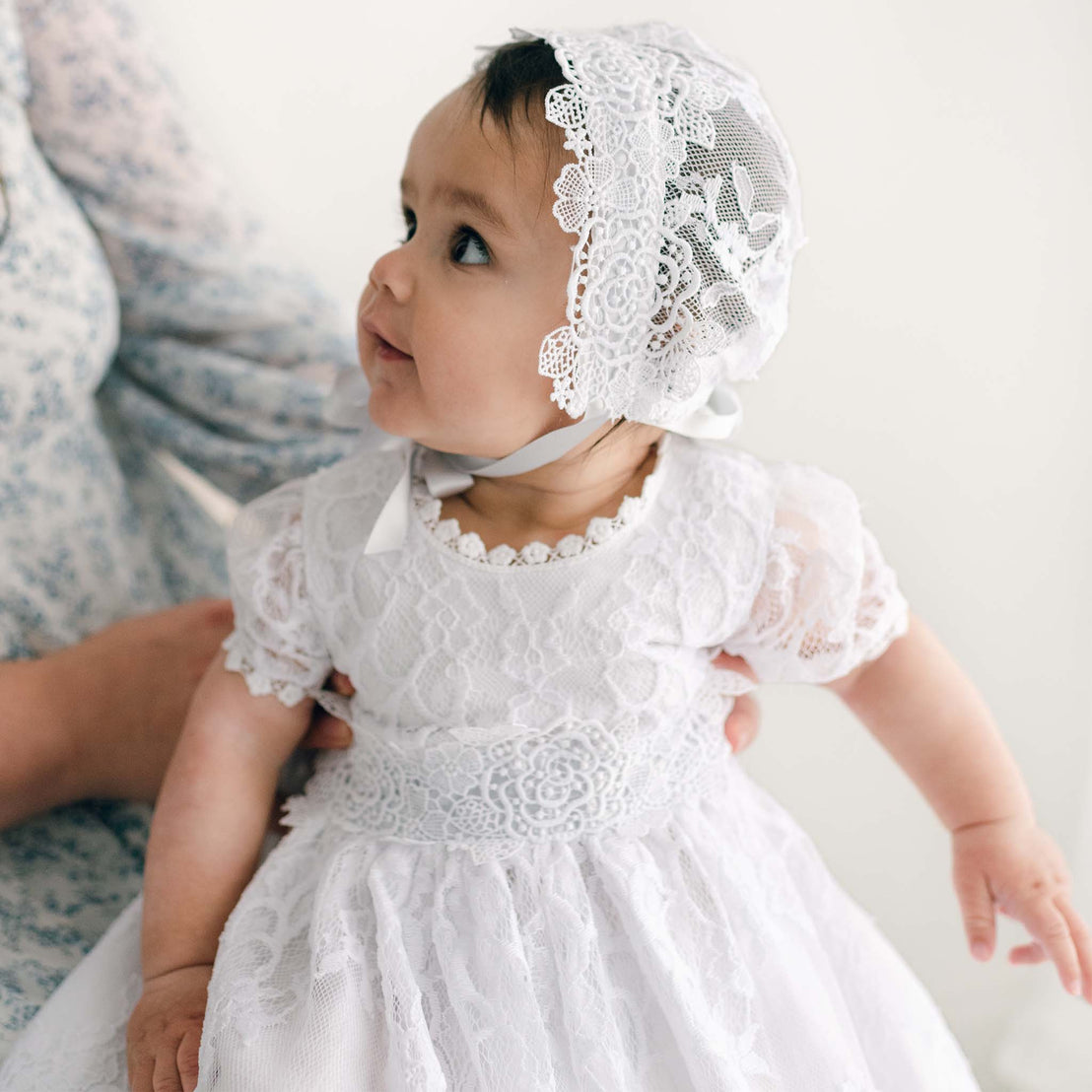 A baby in an Olivia Christening Gown & Bonnet looks up during a baptism, held by an adult against a light-colored background.