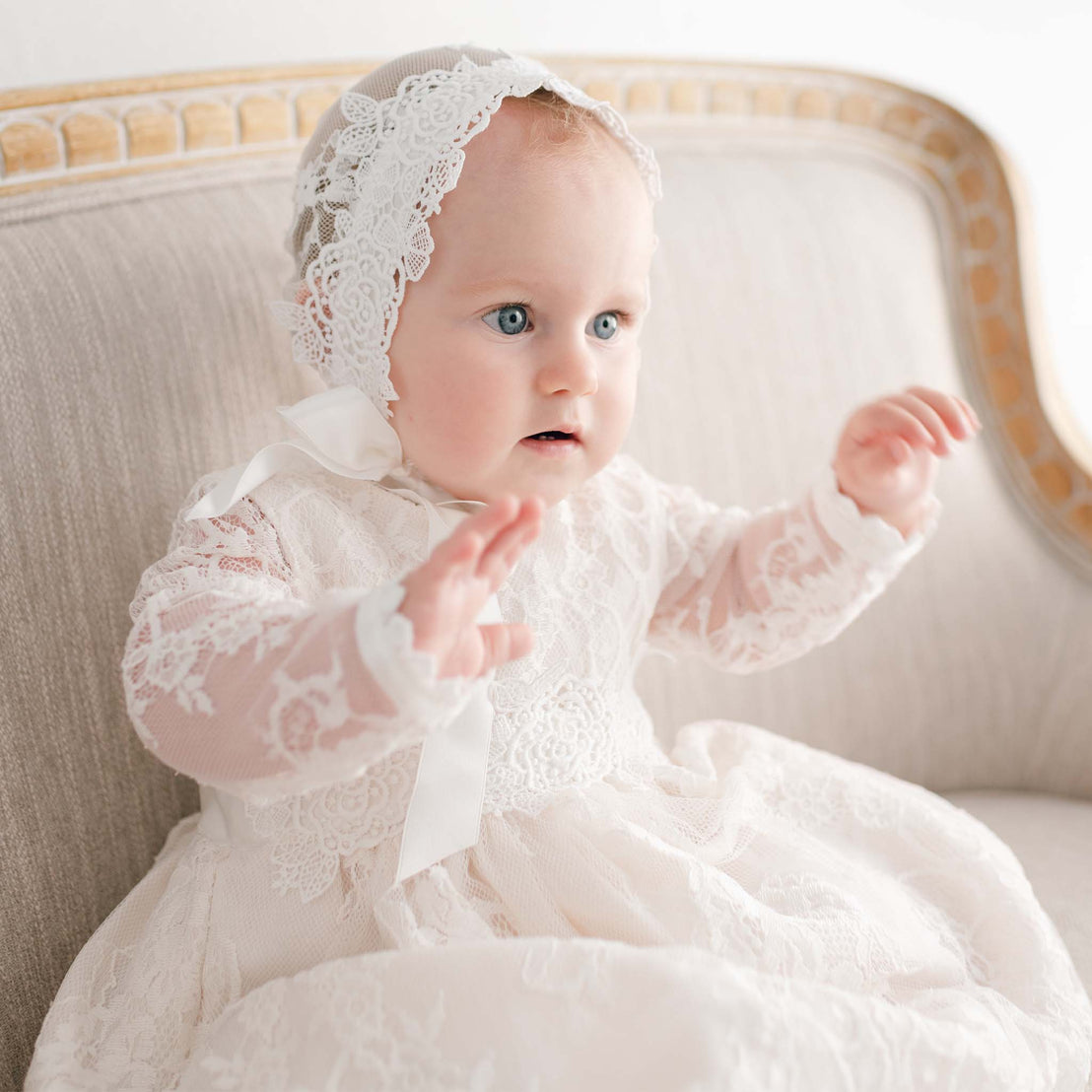 A baby with wide blue eyes wearing a Juliette Christening Gown & Bonnet sits on an upscale couch, looking curiously off to the side.