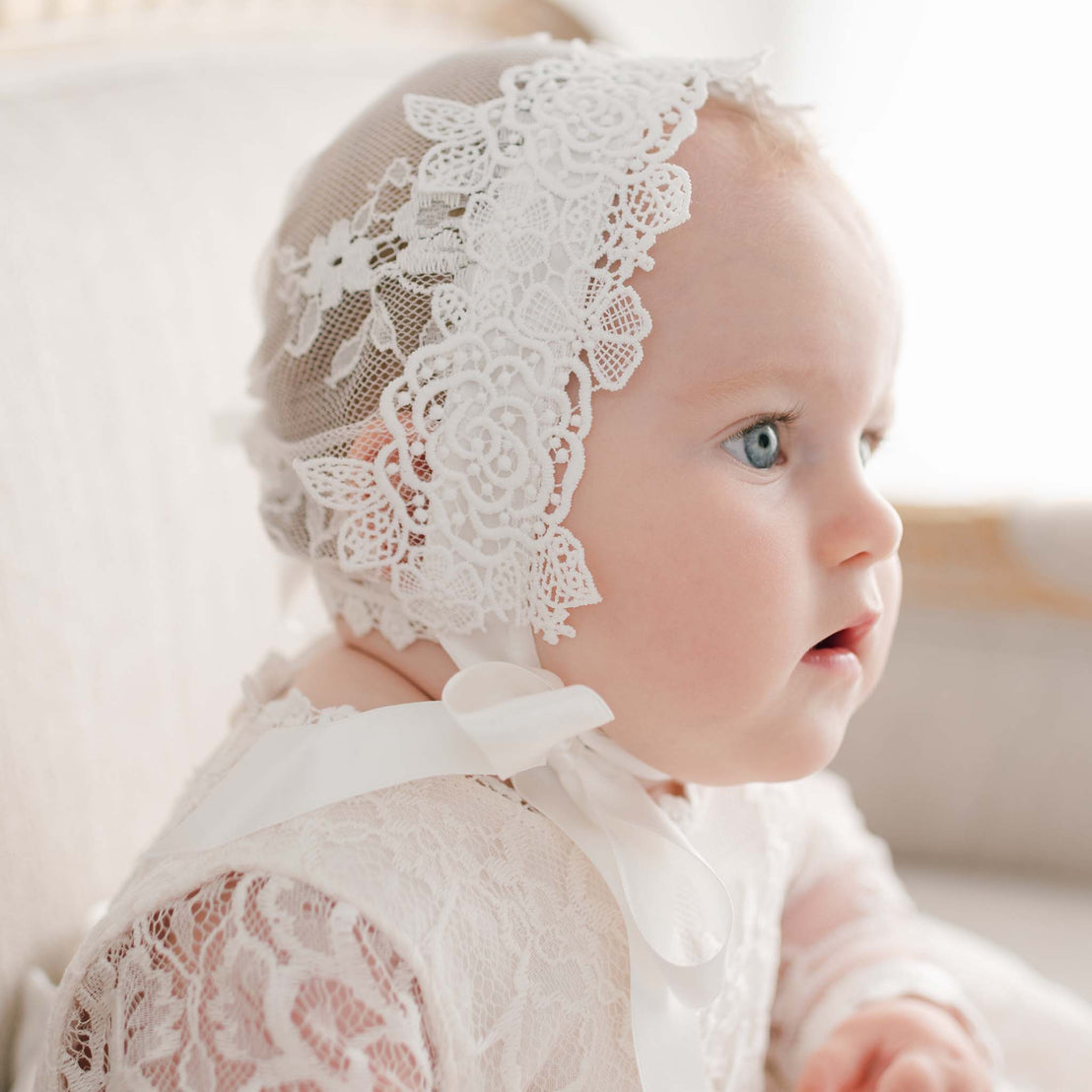 A baby with blue eyes and light skin wearing the Juliette Christening Gown & Bonnet, gazing to the side in a softly lit room.