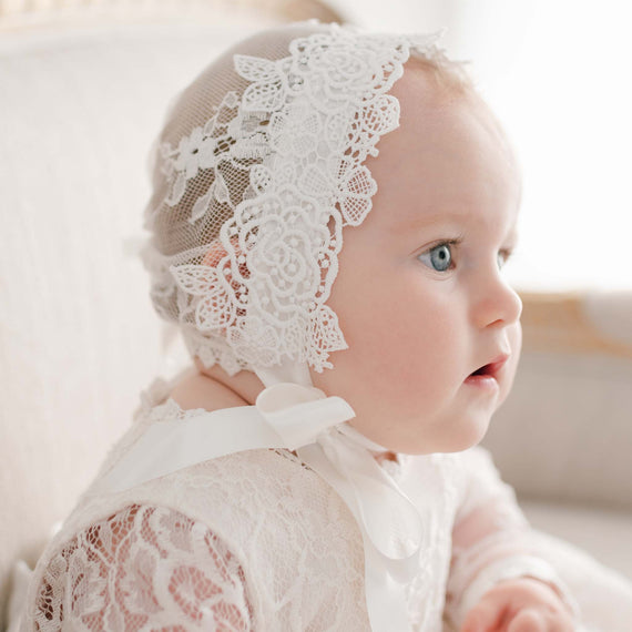 A close-up photo of a baby wearing a delicate white lace dress and a matching Juliette Lace Bonnet, looking off to the side with bright blue eyes. Light filters softly through a background window. The dress