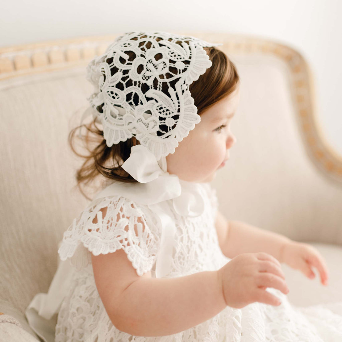 Baby girl sitting on a chair and wearing the Lola Christening Gown and Bonnet.
