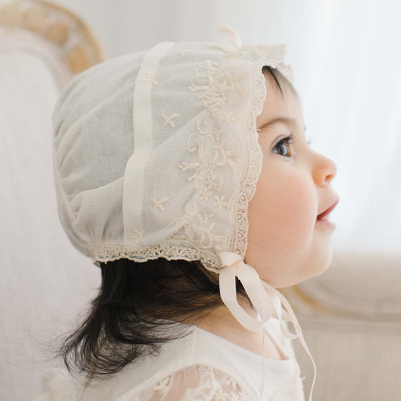 A young child in a delicate Jessica Linen Bonnet looks upward with a gentle expression, highlighted by soft, natural light. The bonnet is adorned with intricate embroidery and secured with a ribbon.