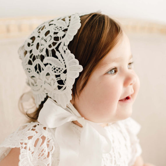 A toddler with fair skin and brown hair smiles gently, wearing the Lola Lace Bonnet and a white dress with lace details for a vintage baptism. The soft, natural light enhances the serene ambiance.