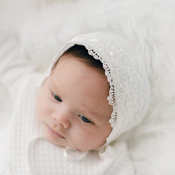 A newborn baby wearing a Madeline Quilted Newborn Bonnet, an heirloom piece, lies on a soft, fluffy white blanket, looking thoughtfully to the side.