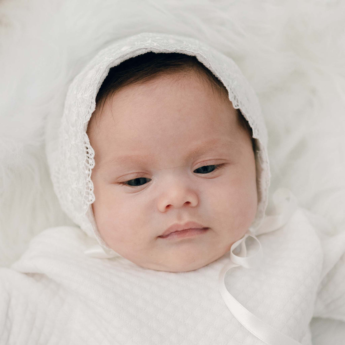 A serene baby with a slight frown, wrapped in a Madeline Quilted Newborn Bonnet, against a fluffy white backdrop.