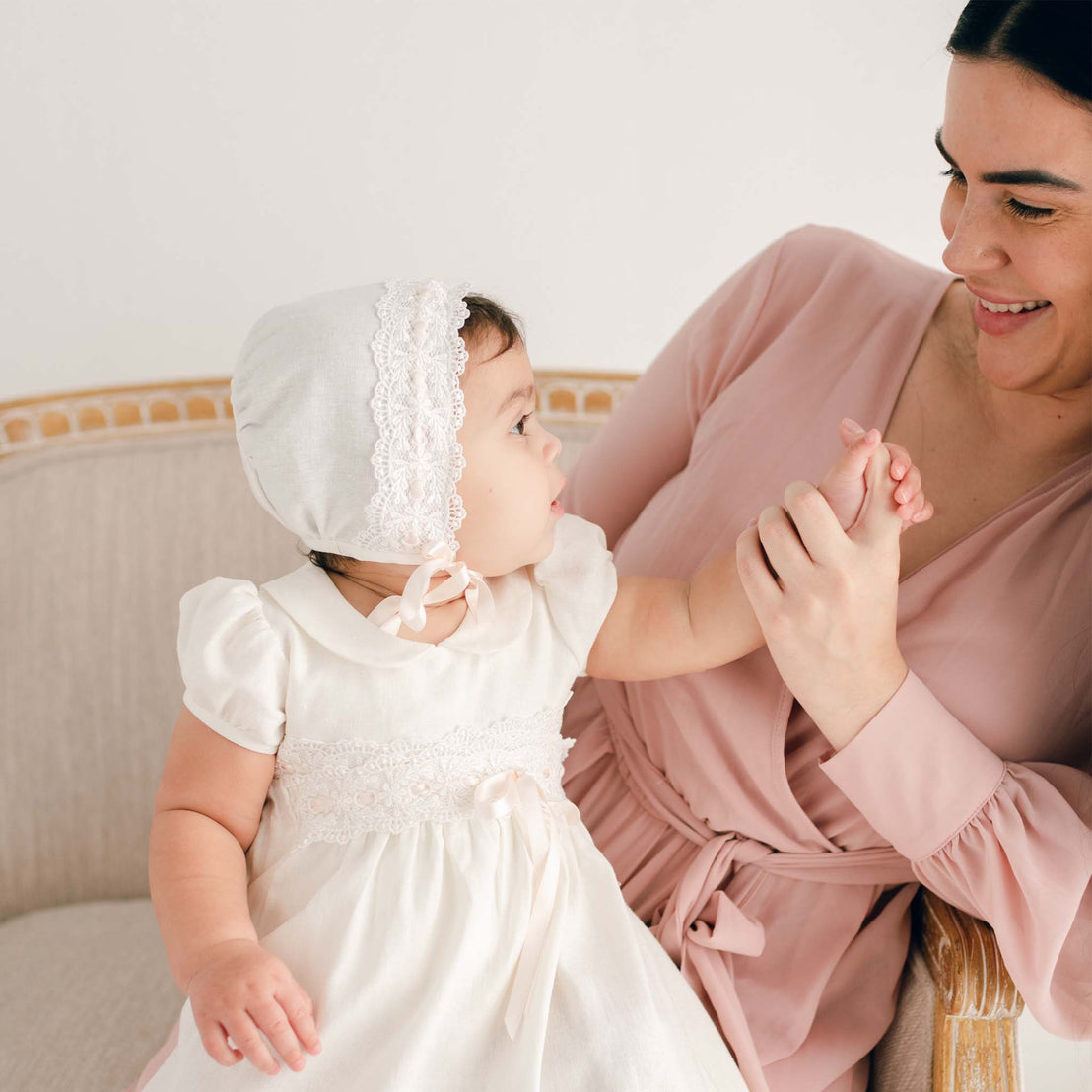 A mother in a pink dress smiles at her baby girl, who is dressed in a boutique white lace christening gown and Emma Bonnet, as they sit together on a beige sofa.