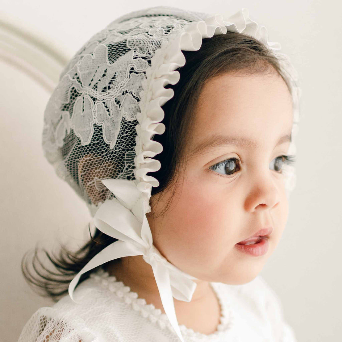 Close up of a baby girl wearing the Victoria Lace Bonnet. The bonnet is made in a fitted style with ivory floral lace, ivory silk ribbon ties and ruffles.