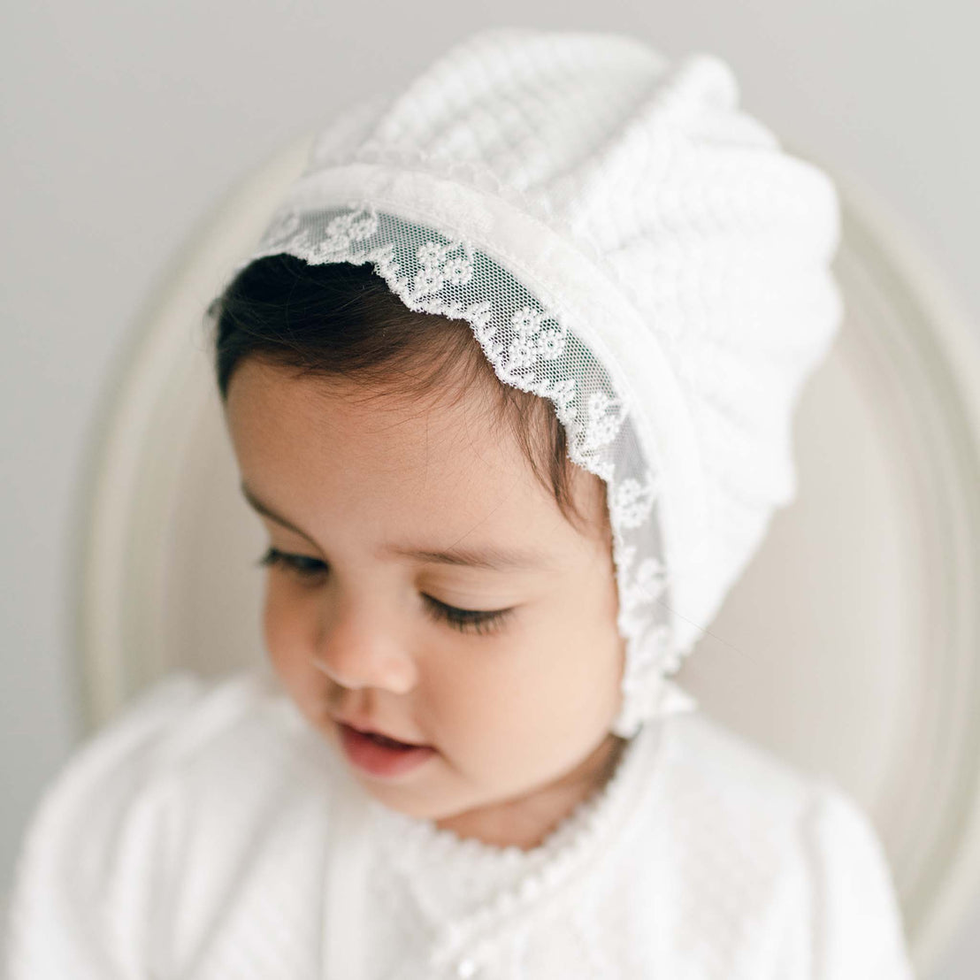 Baby girl wearing the Emily Quilted Cotton Bonnet made from a soft quilted cotton in a shade of white. The bonnet is also adorned with light ivory floral lace trim and white velvet ribbon.
