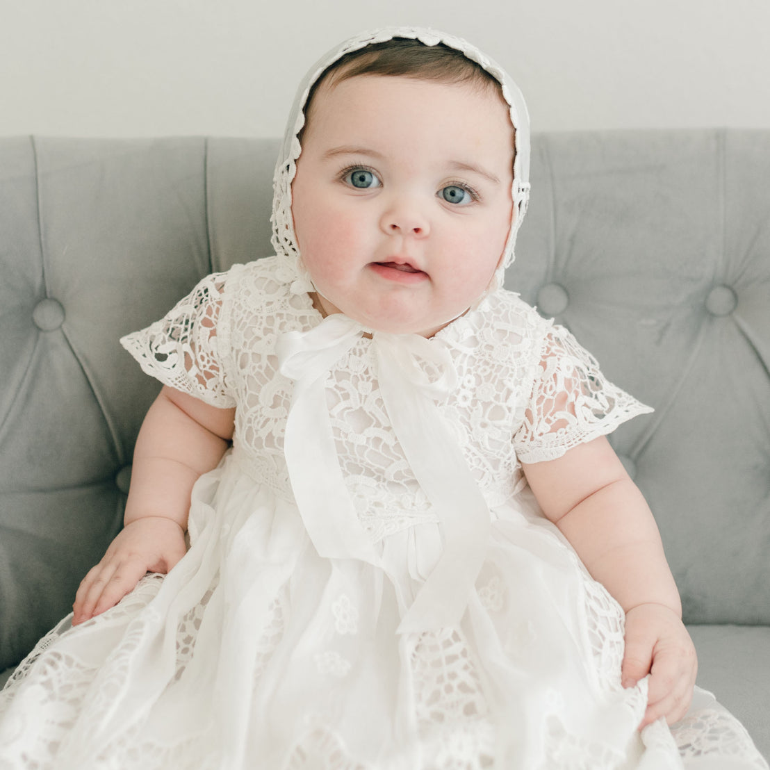 A baby with big blue eyes wearing a Grace Christening Gown & Bonnet sits on a gray sofa, looking directly at the camera with a curious expression.