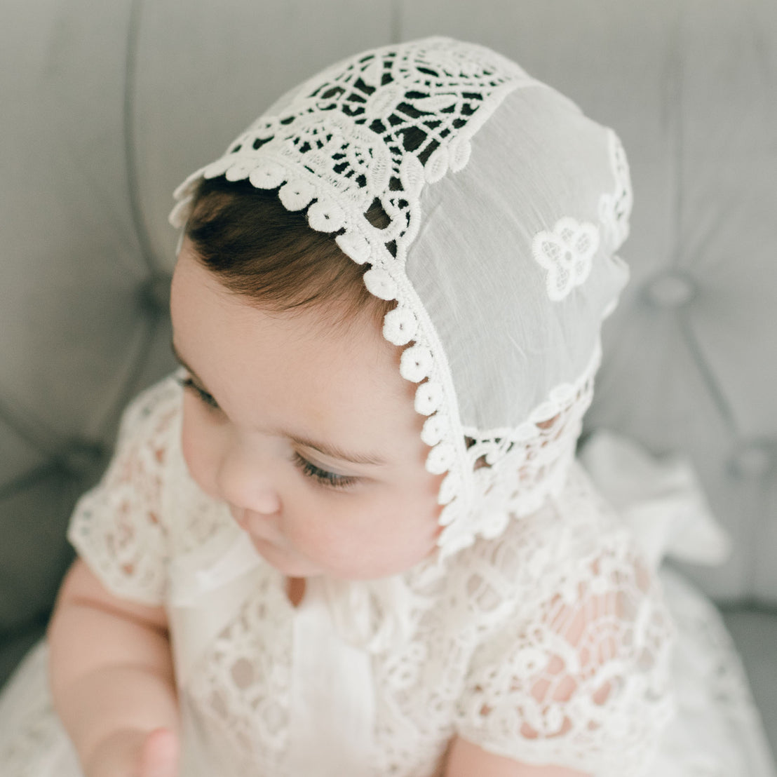A serene baby wearing the Grace Christening Gown & Bonnet, with a Venice inset and a silk sash tie, sitting on a grey textured background, gazing down thoughtfully.