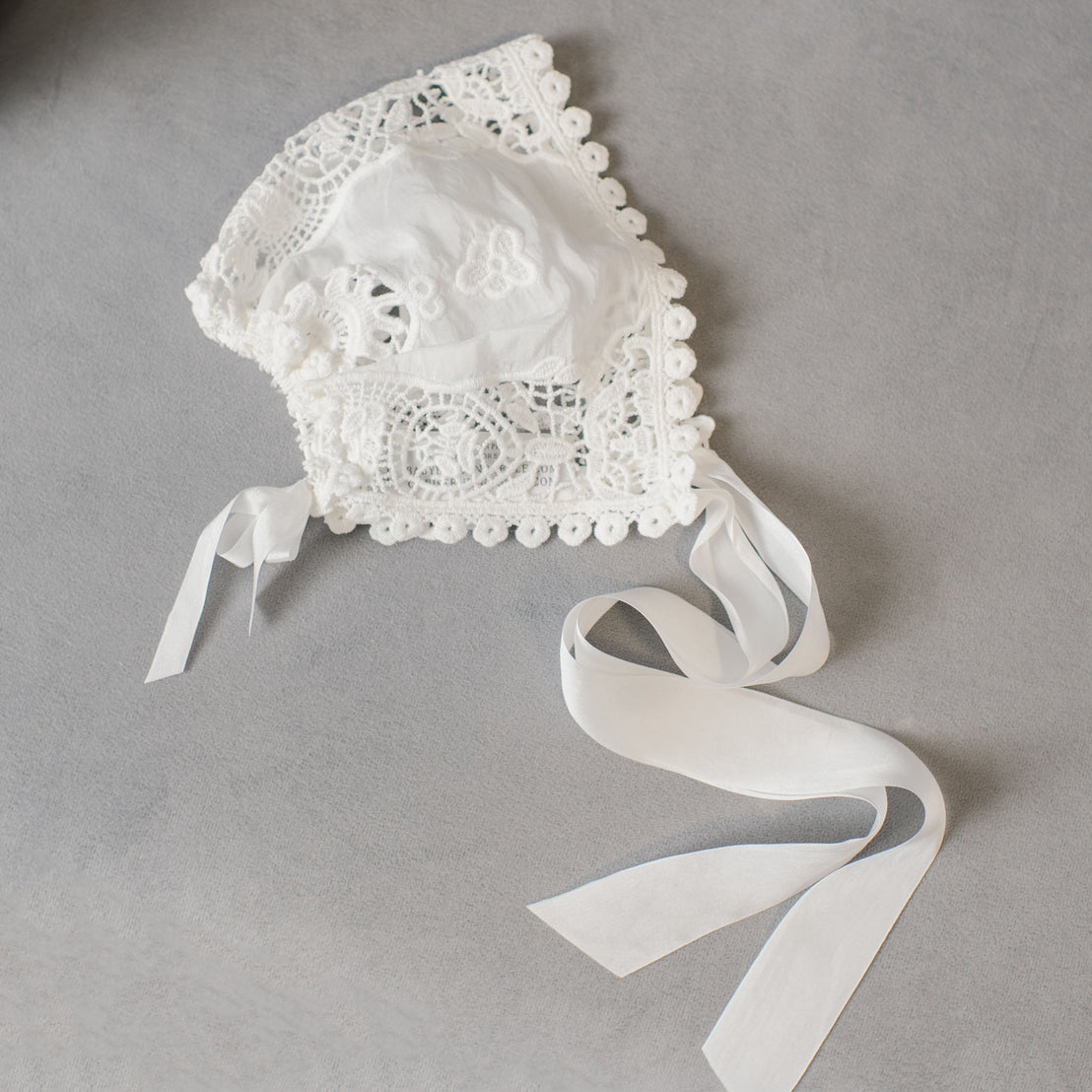 A delicate Grace Christening Bonnet with silk sash tie photographed on a gray background, showcasing intricate patterns and floral designs.