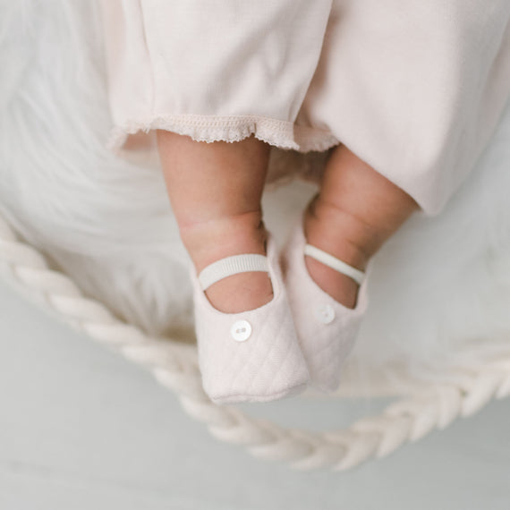 A close-up image of a baby's feet wearing Ava Quilted Booties with button details, the feet are gently resting inside a woven basket lined with white fur rug.