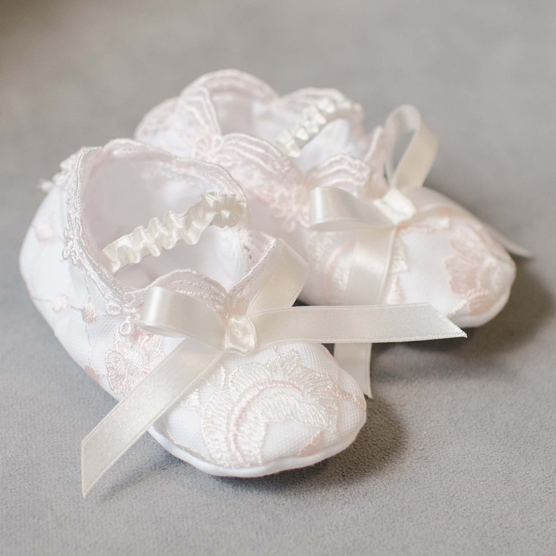 A pair of delicate white Joli Booties adorned with lace and satin ribbons, perfect for a baptism, displayed on a soft gray background in an upscale boutique.
